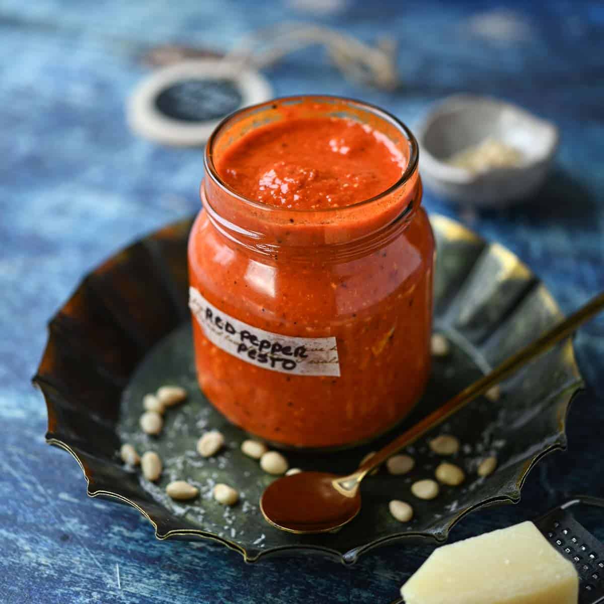 Clear jar of roasted red pepper pesto with a spoon on a blue surface.