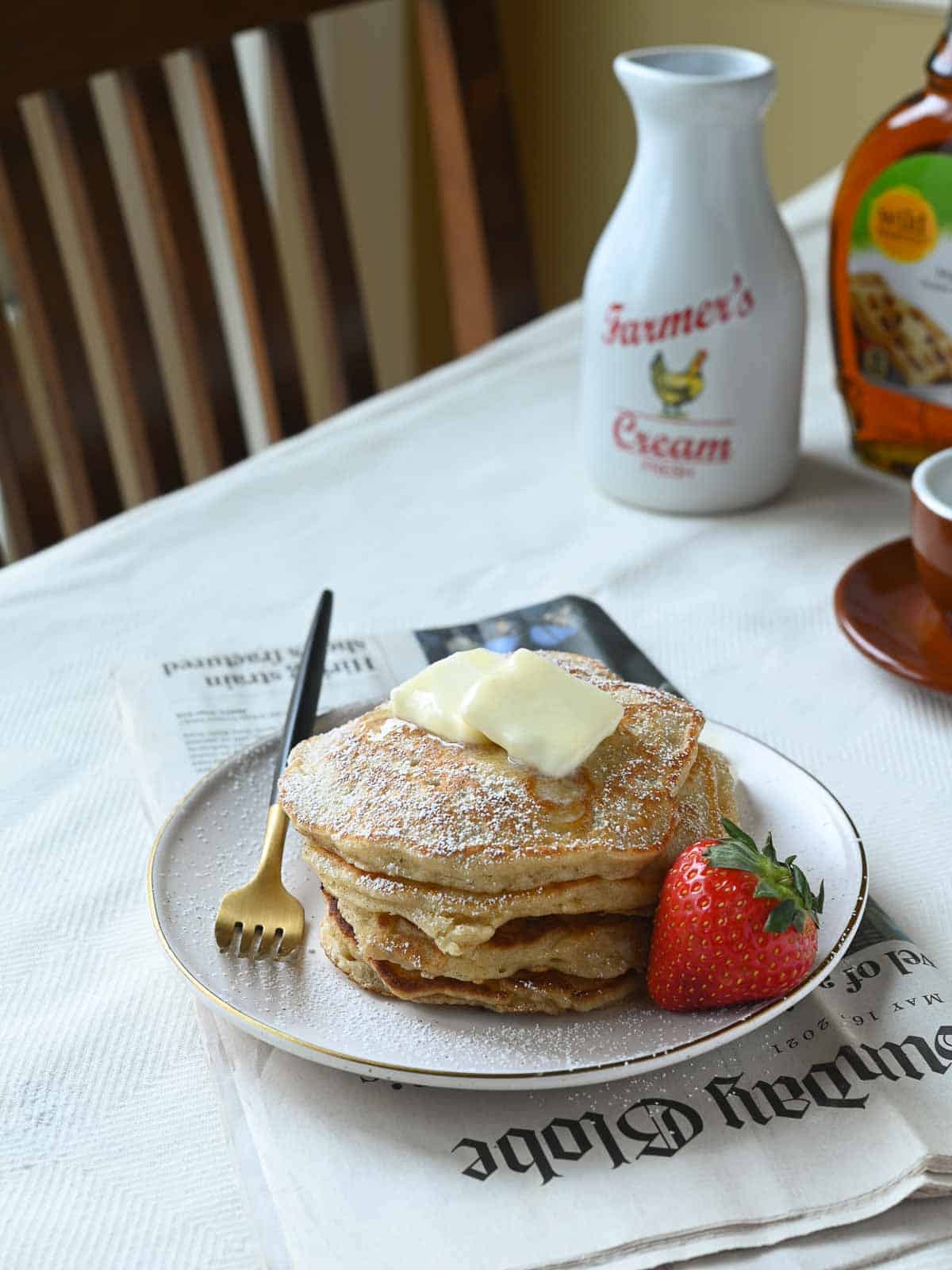 Pancakes on a kitchen table with a strawberry to the side and the morning paper under the plate.