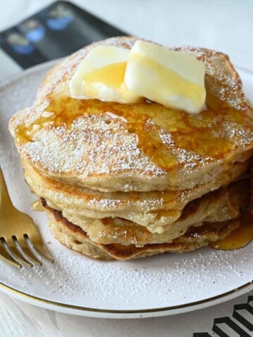 Close-up of pancakes with butter and maple syrup on a beige plate with a fork to the left.