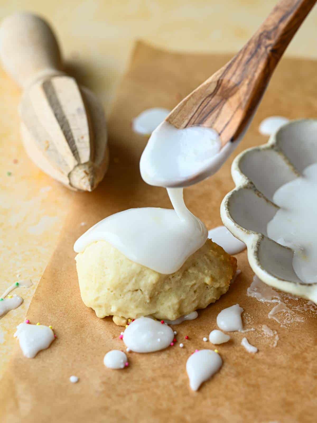 Lemon icing being dripped by a spoon over a cookie on top of brown parchment paper.