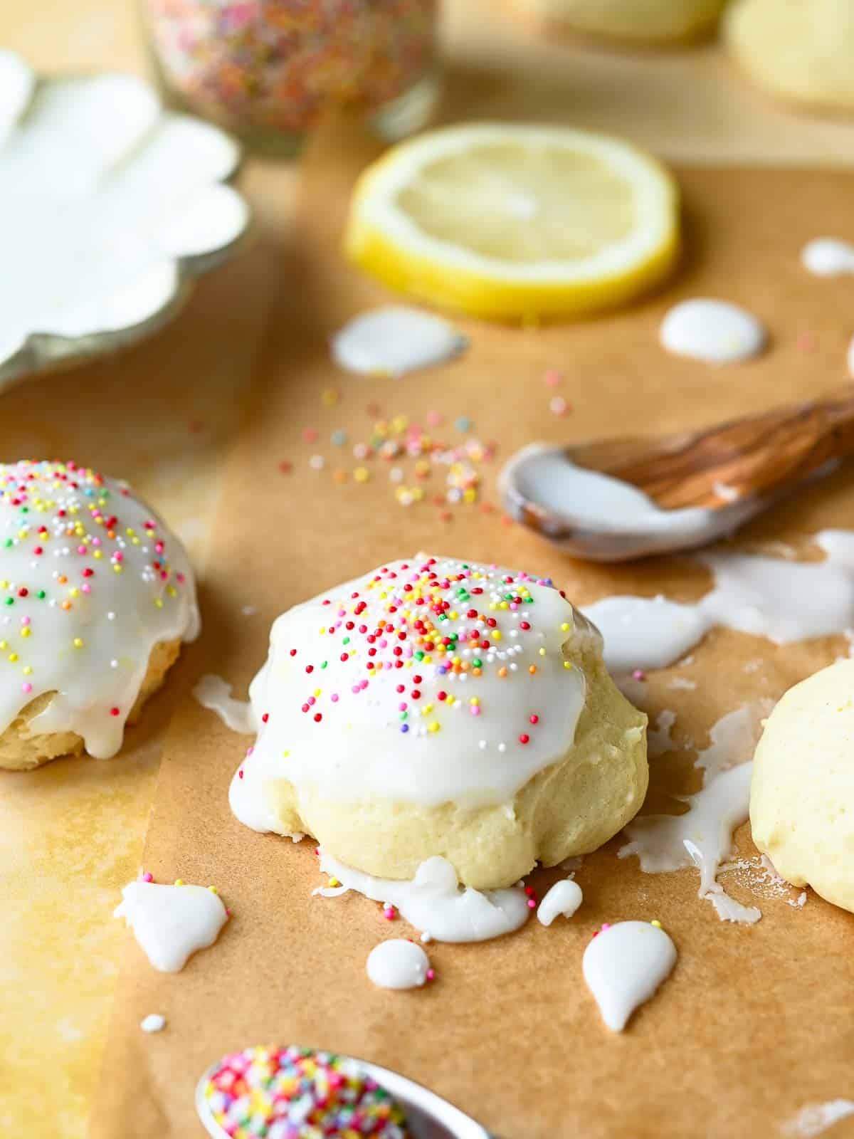 Italian lemon drop cookies with icing and sprinkles on parchment paper with lemons and sprinkles in the background.