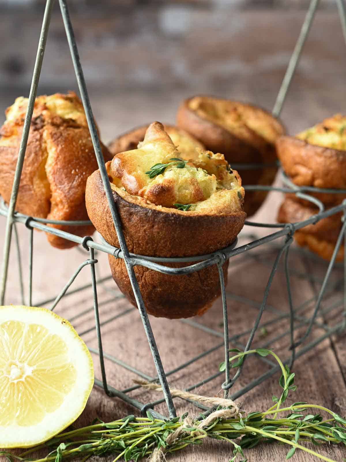 Lemon popovers are suspended in a wire rack with thyme and lemons in the forefront.