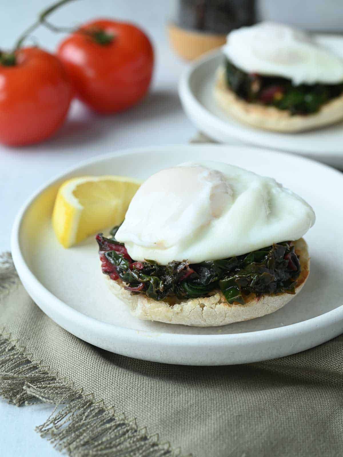 Two white plates with English muffins, swiss chard, and poached eggs on them.
