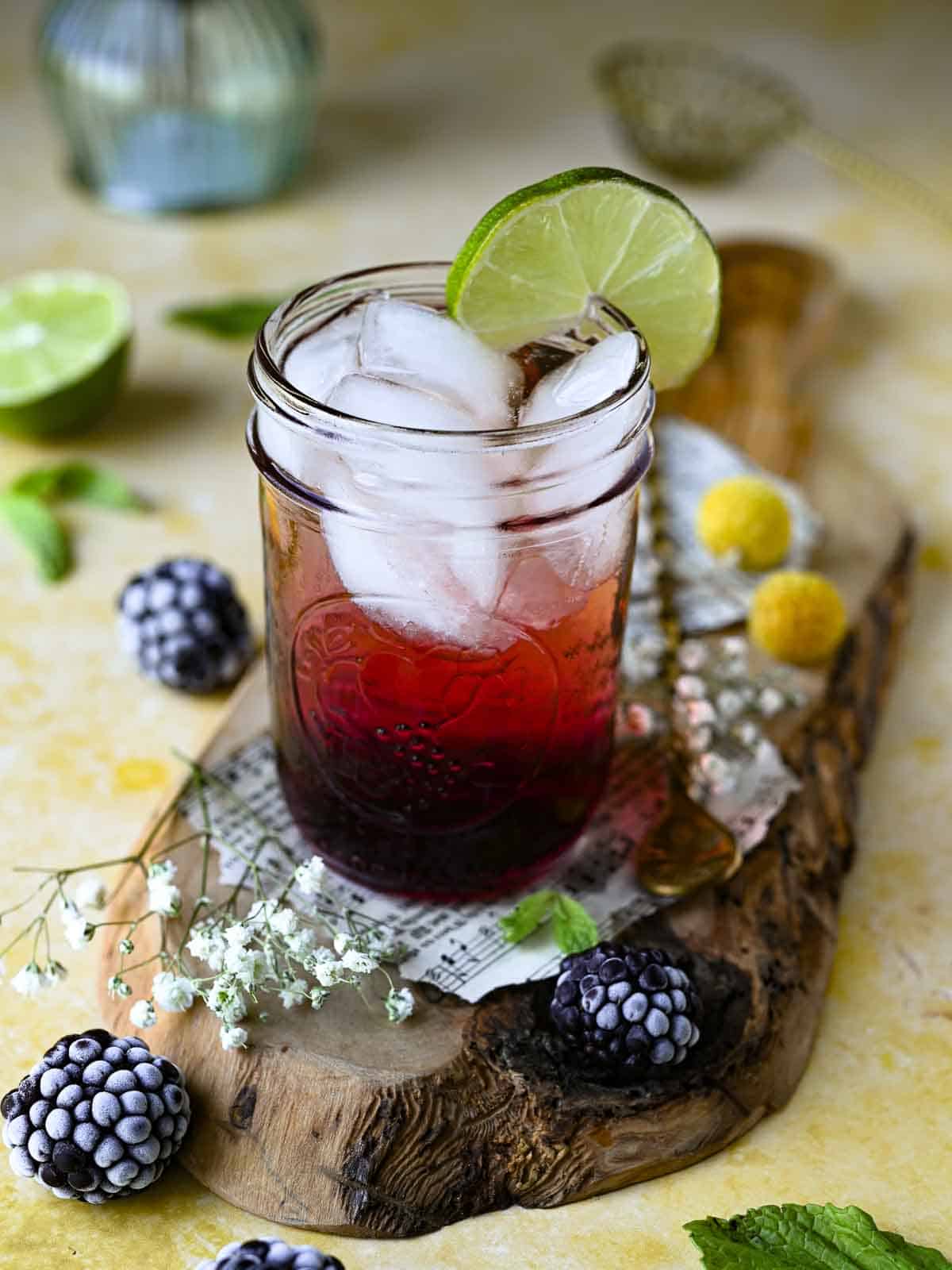 Blackberry soda in a clear glass on wood with blackberries and flowers around it.