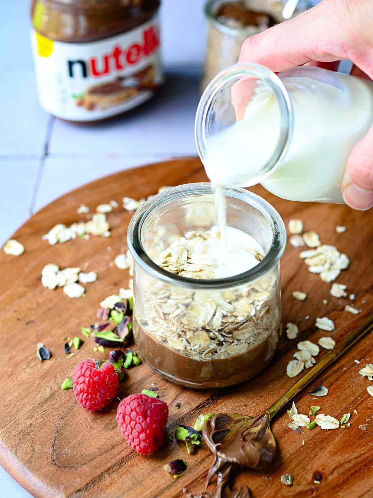 Milk is poured into a jar of oats and Nutella on wood.