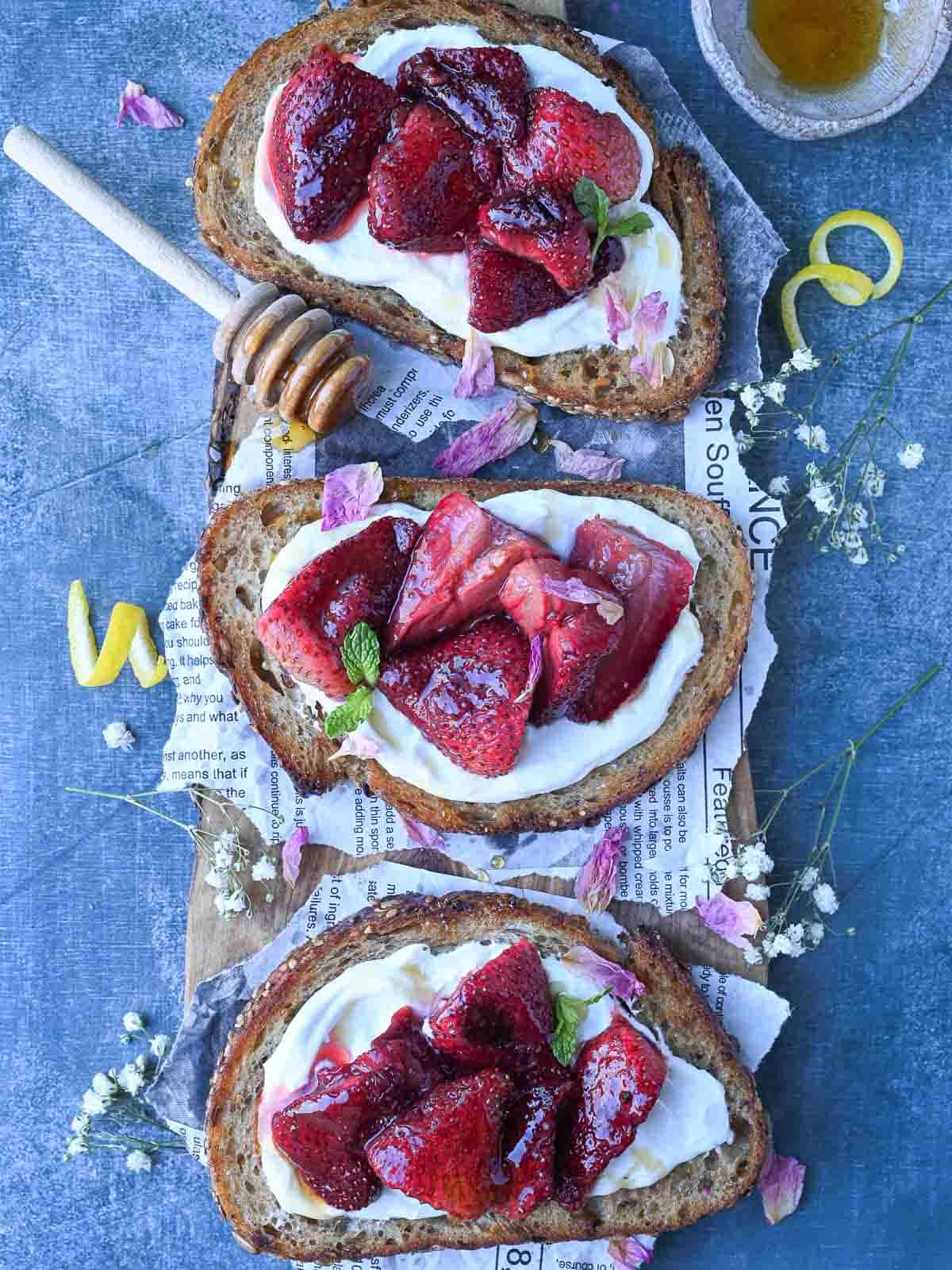 Overhead of three labneh toasts with strawberries on a blue surface.