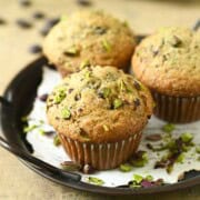 Three pistachio muffins on a black tray with scattered pistachios.