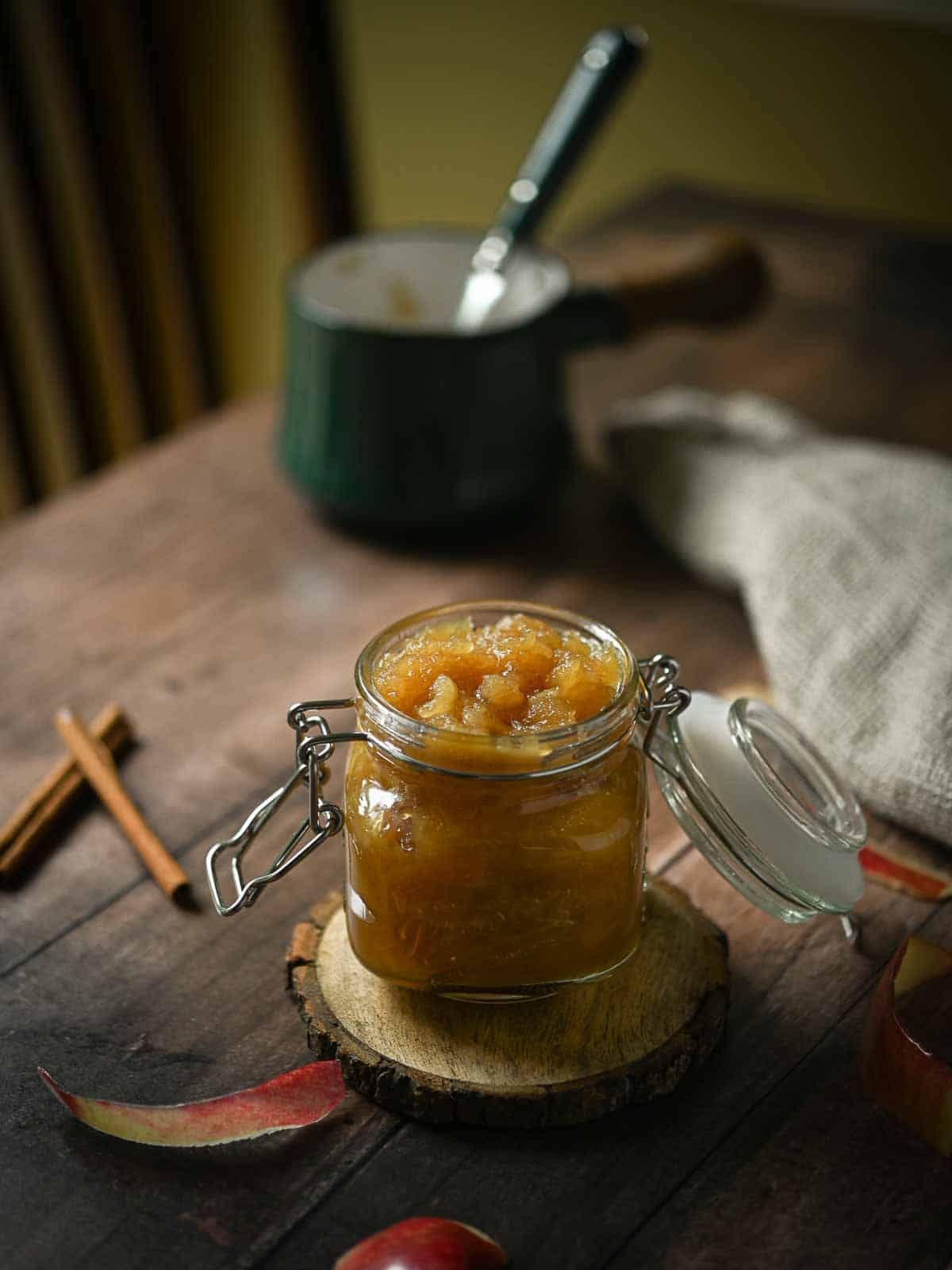 A moody shot of Honeycrisp applesauce in a clear jar on a wooden table with a pot in the background.