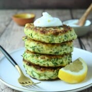 Stacked zucchini pancakes on a beige plate with a lemon and fork on the side.