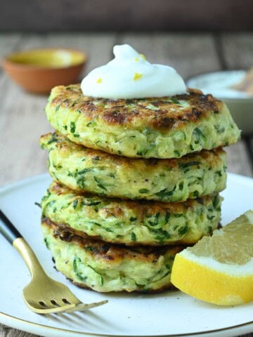 Stacked zucchini pancakes on a beige plate with a lemon and fork on the side.