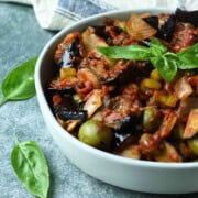 Close-up of eggplant caponata in a white bowl with basil leaves on top.