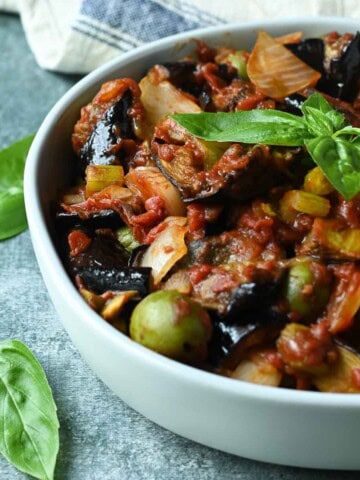 Close-up of eggplant caponata in a white bowl with basil leaves on top.