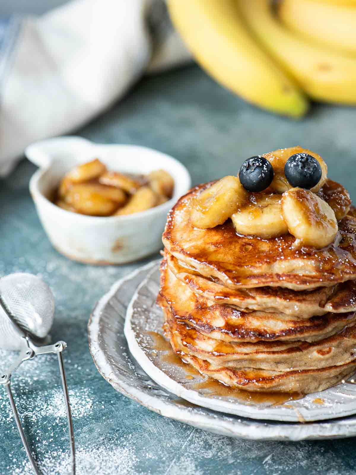 A stack of pancakes with bananas on top placed on grey plate over a blue surface.