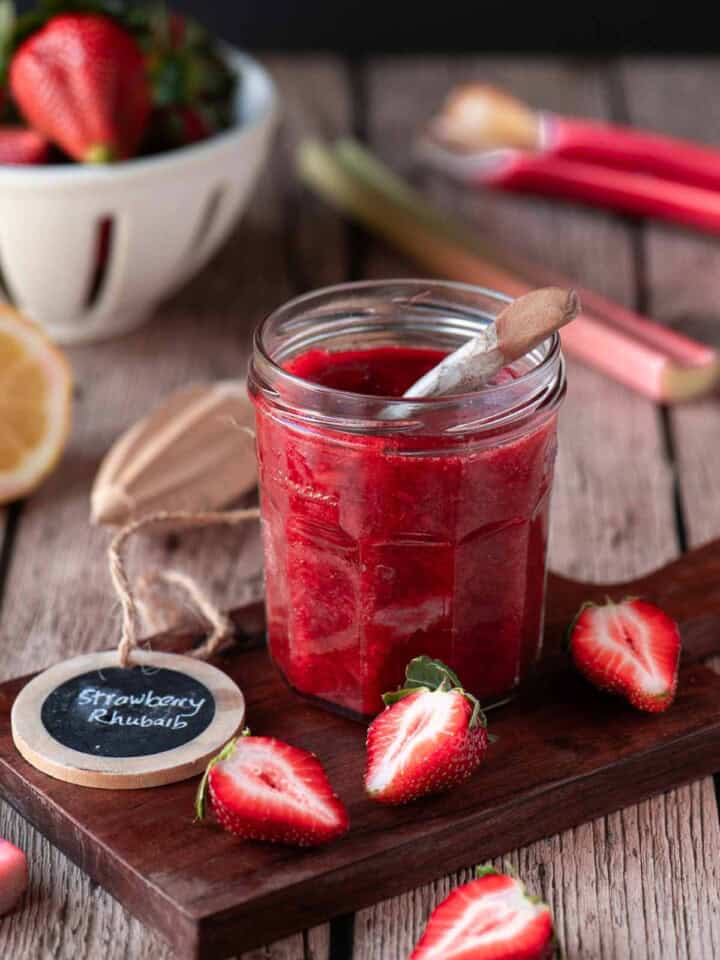Strawberry rhubarb jam in a clear jar on a wood surface with fruit around it.