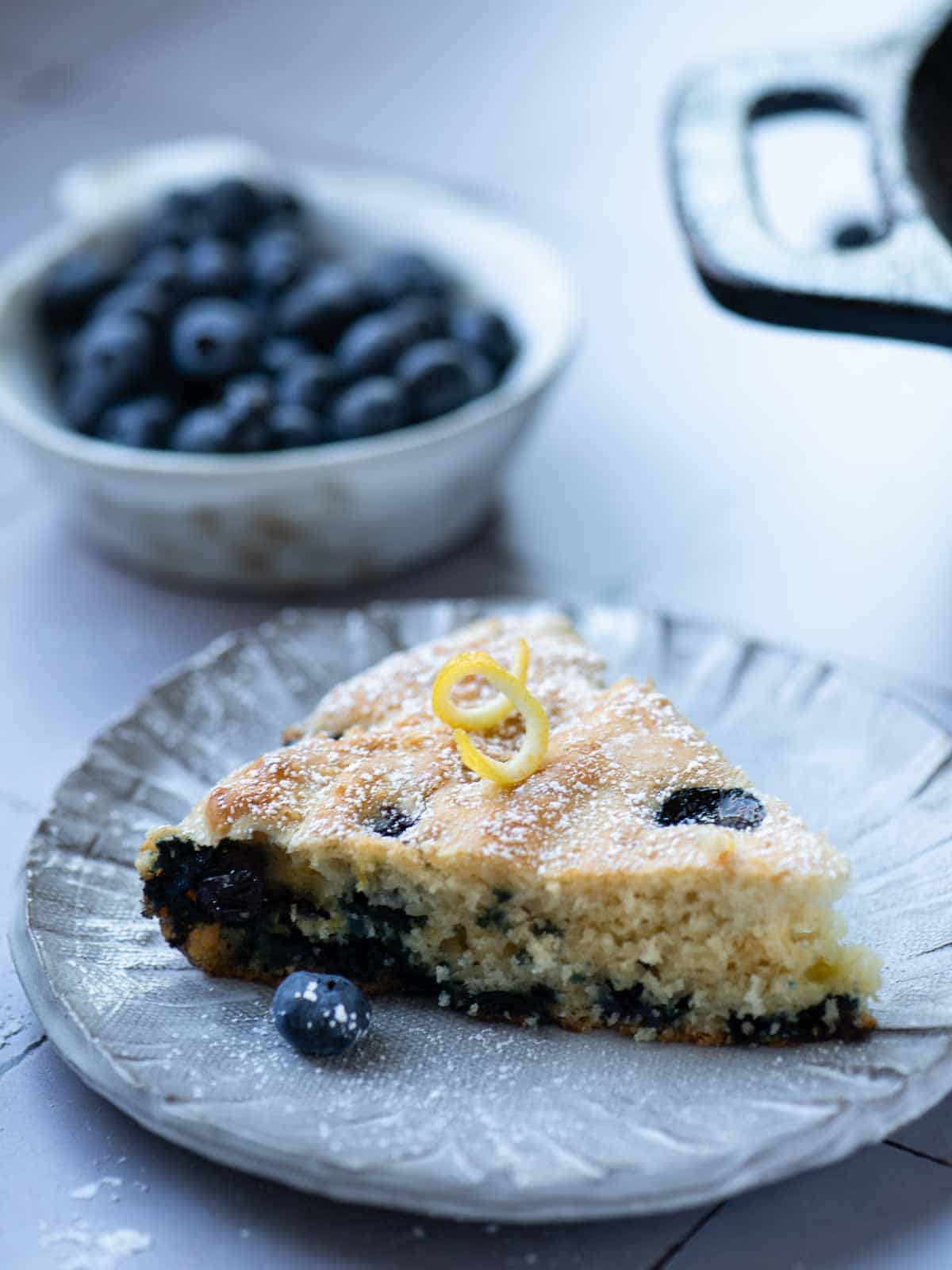 A slice of blueberry skillet pancake on a grey plate with blueberries in the background.