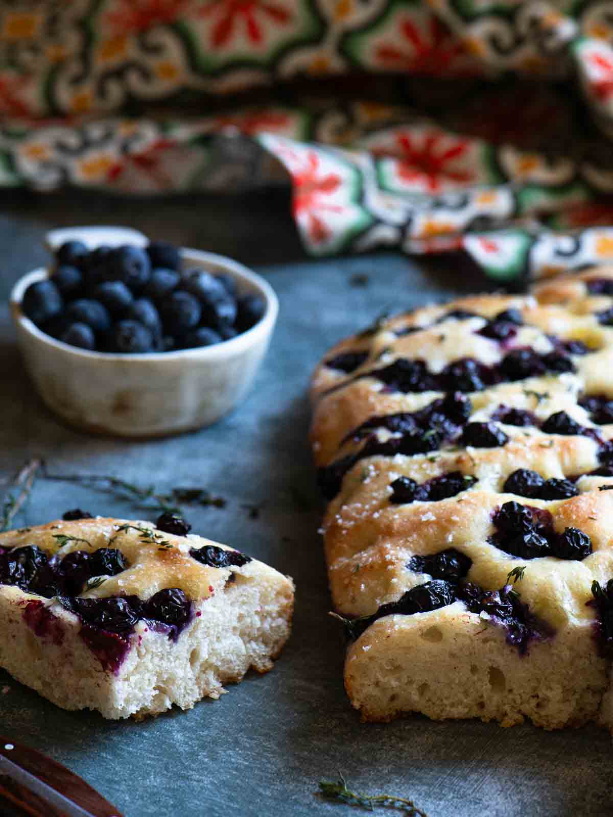 A slice of blueberry focaccia on a grey surface with blueberries in the background.