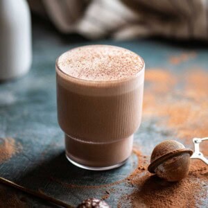 Chocolate smoothie in a clear glass on a blue surface with cocoa powder around it.