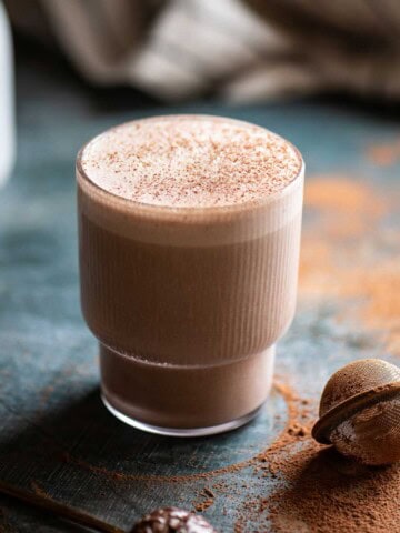 Chocolate smoothie in a clear glass on a blue surface with cocoa powder around it.