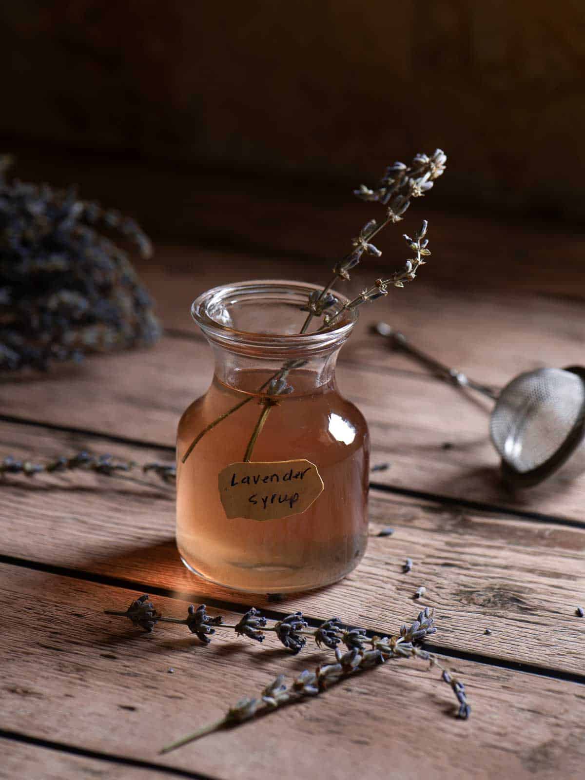Lavender syrup in a clear container on a wood surface with lavender buds in it.