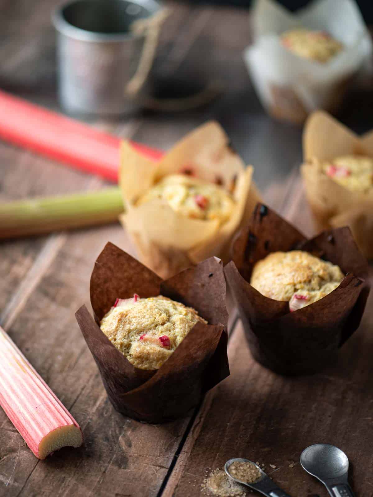 Five rhubarb muffins on a wooden surface with rhubarb stalks next to it.
