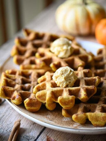 A platter of waffles on a wooden table with pumpkins in the background.