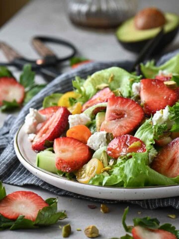 Close up of a strawberry salad with lettuce, tomatoes, goat cheese on a beige surface.