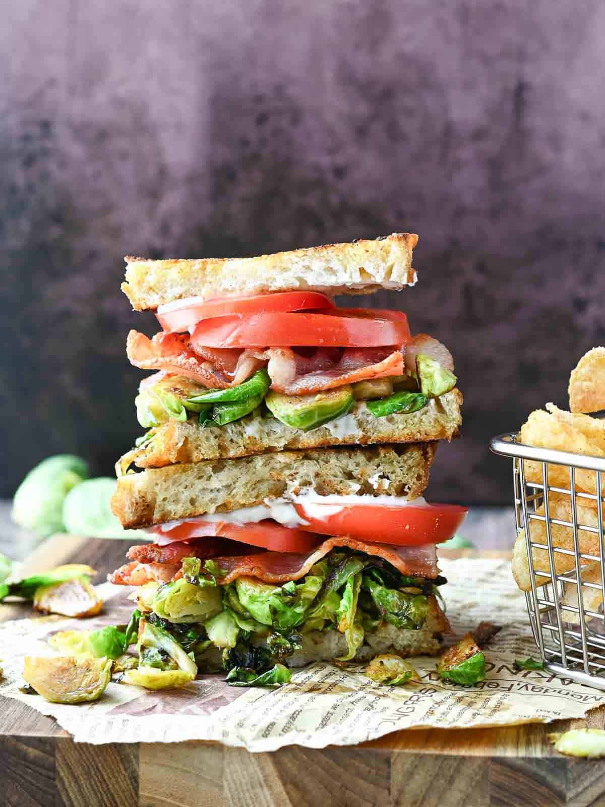 Two BLTs stacked on top of each other with brussel sprouts and potato chips.