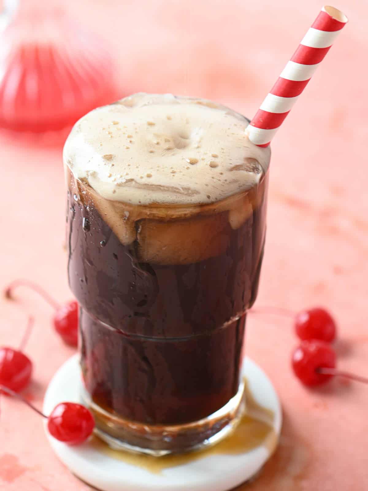 A cherry americola drink in a clear glass with a straw on a pink surface.