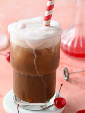 A cherry americola drink in a clear glass with a straw on a pink surface.