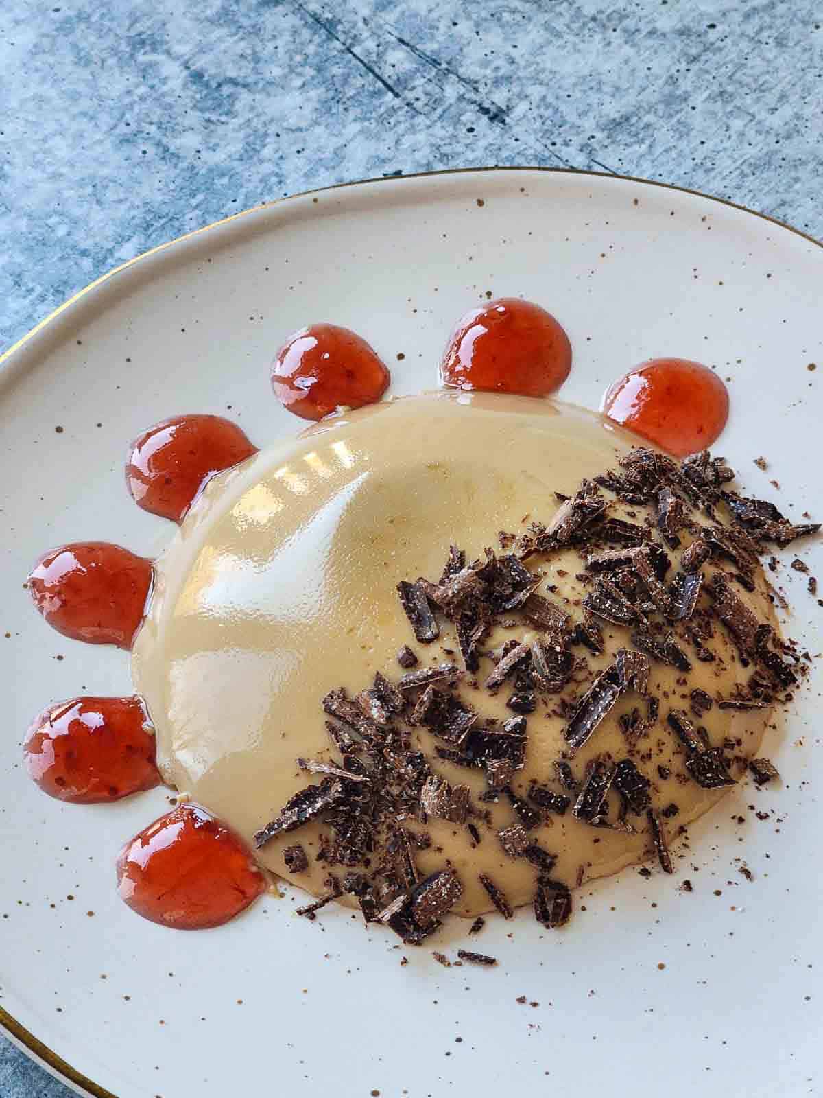 Panna cotta on a beige plate with raspberry swirls and shaved chocolate.