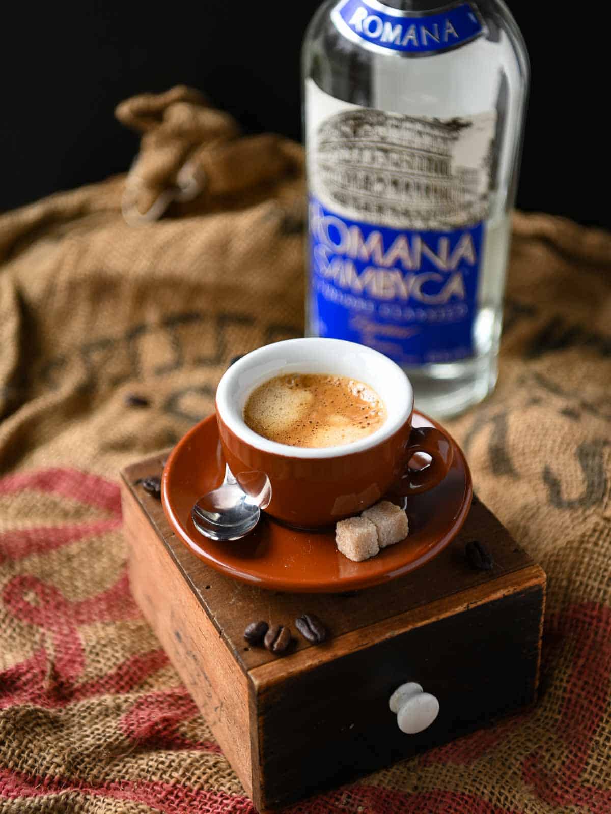 A cup of espresso elevated on a wooden box with a bottle of sambuca in the background.