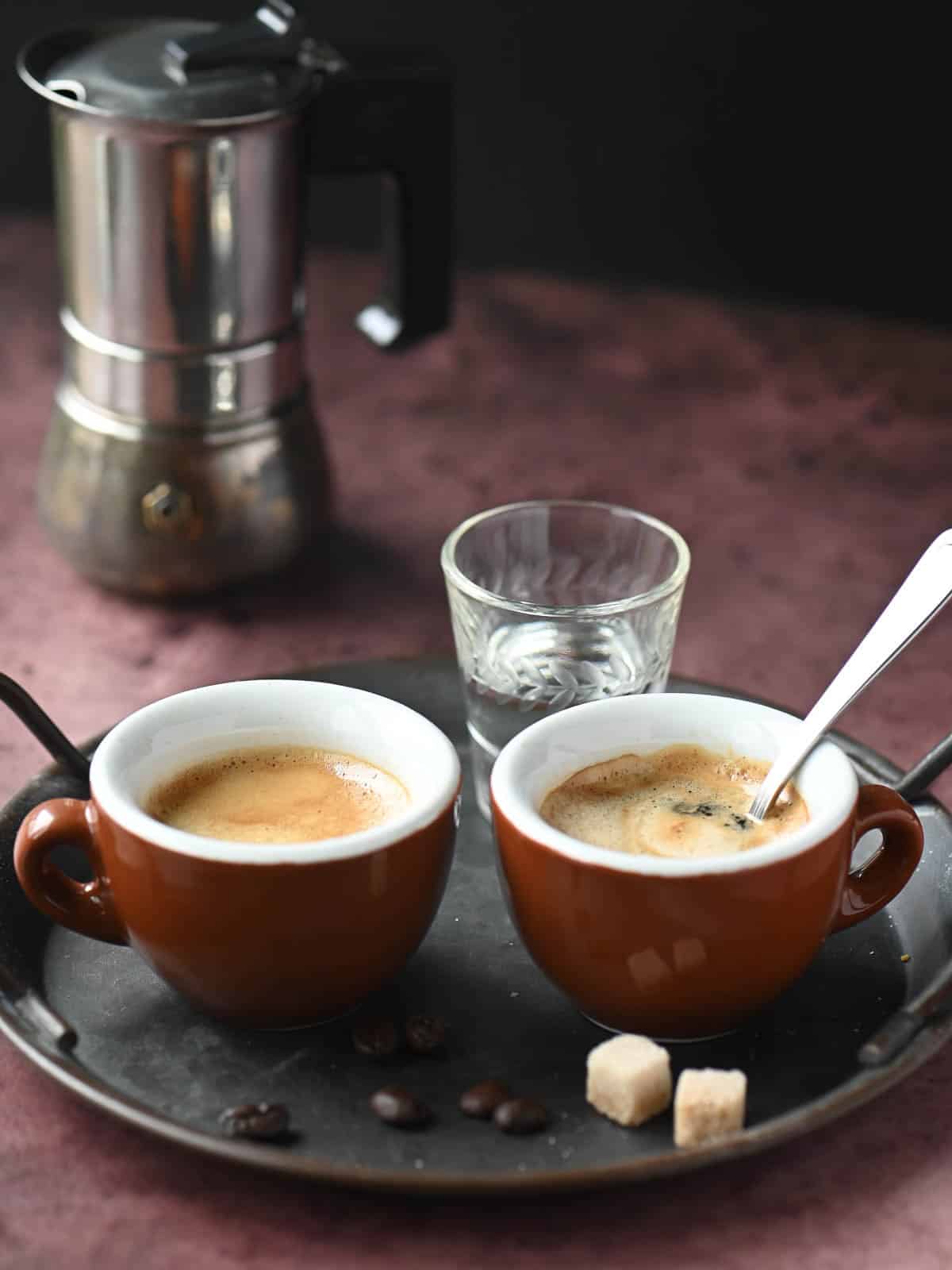 Two cups of espresso on a metal tray with a shot of liquor on the side.