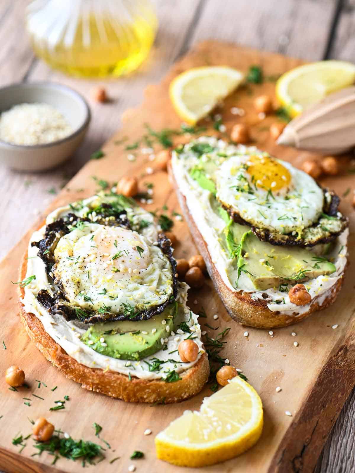 Two hummus toasts on a cutting board with eggs and avocado and lemon slices.