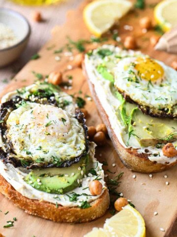 Two hummus toasts on a cutting board with eggs and avocado.
