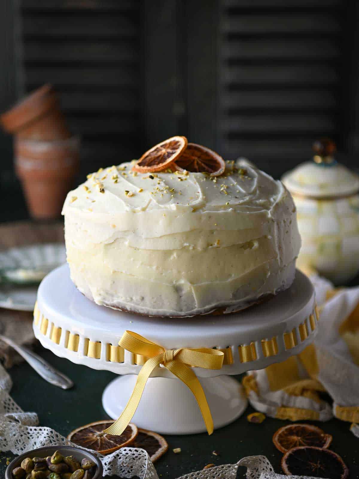 An eye-level view of a whole carrot cake with frosting on a white pedestal with yellow ribbon.