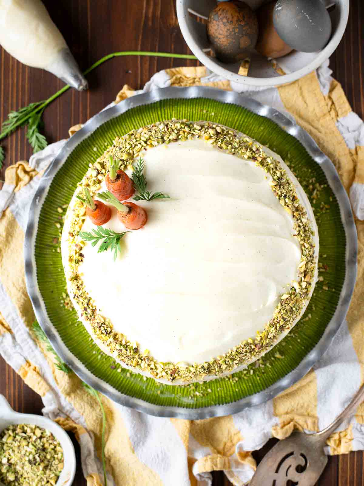 Overhead of a carrot cake with pistachios around the edges on a green plate with 3 carrots.