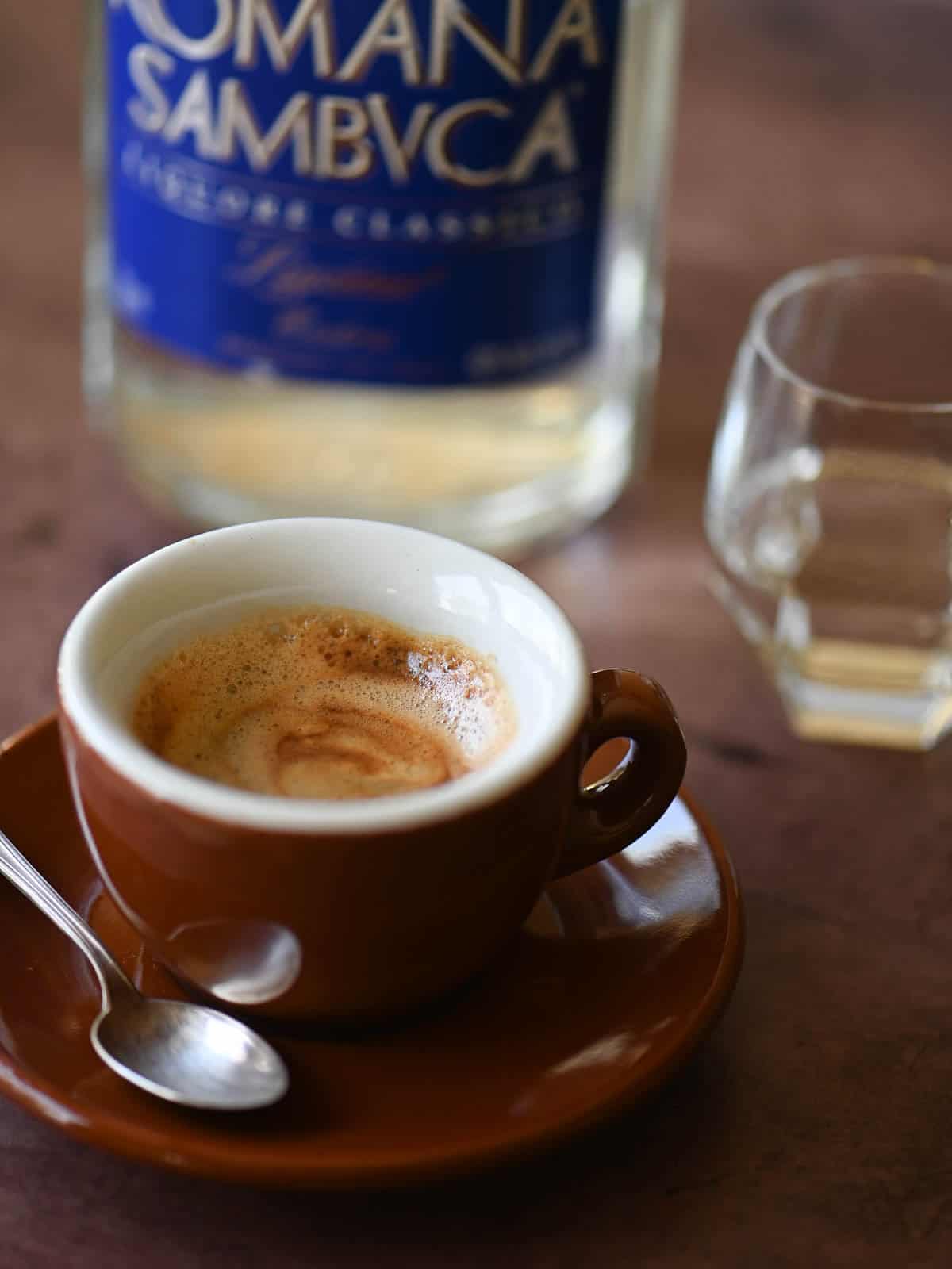 A brown cup of espresso with a bottle of Sambuca in the back.