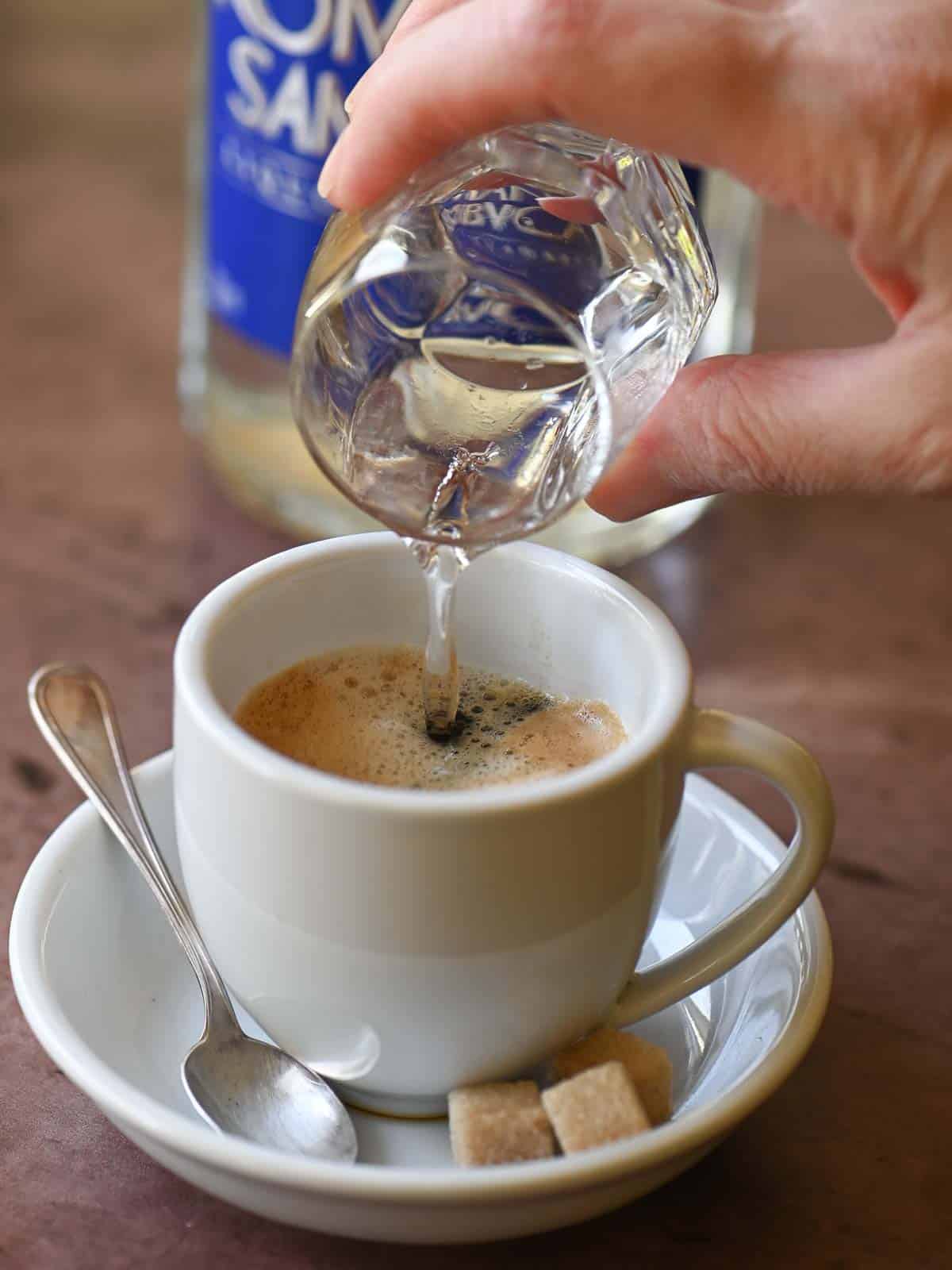 A hand pouring Sambuca into a white cup with espresso.