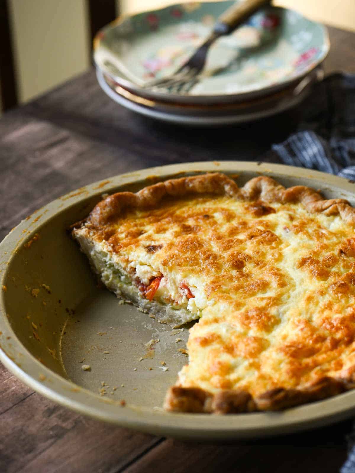 A half-eaten quiche in a metal pan on a wooden table with plates in the background.