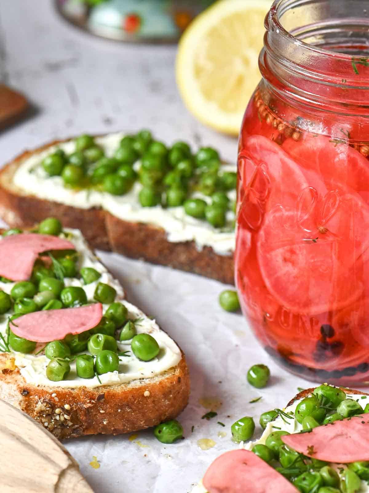 A jar of pickled radishes next to pea toasts on a white board.