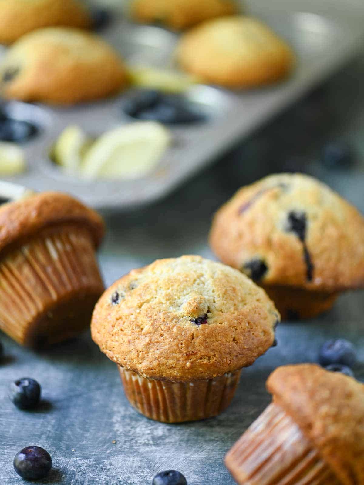 Scattered blueberry muffins in a muffin pan and on a blue surface.