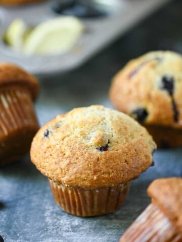 Scattered blueberry muffins in a muffin pan and on a blue surface.