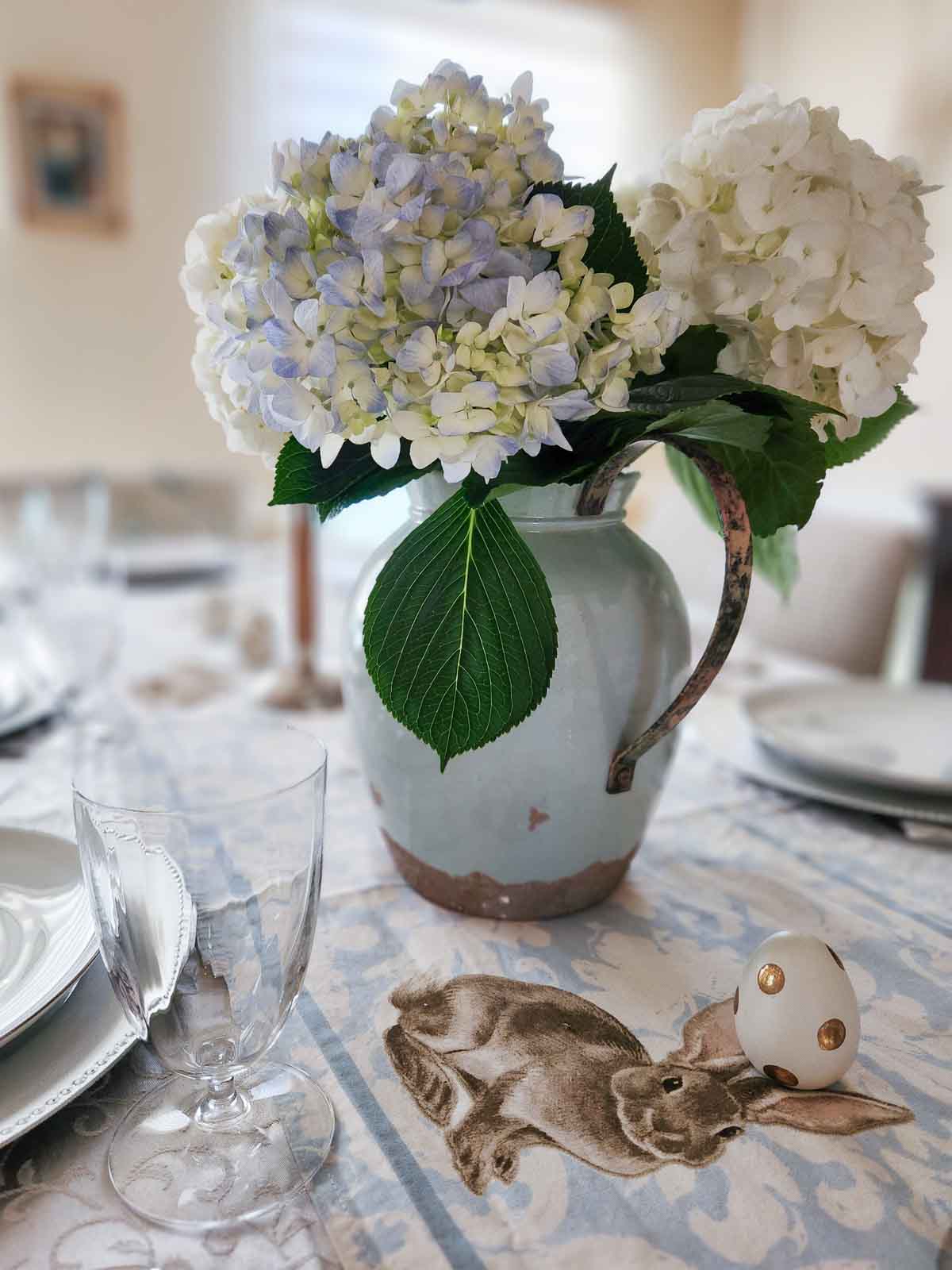 An Easter table decorated with hydrangeas, easter eggs, and a blue rabbit runner.