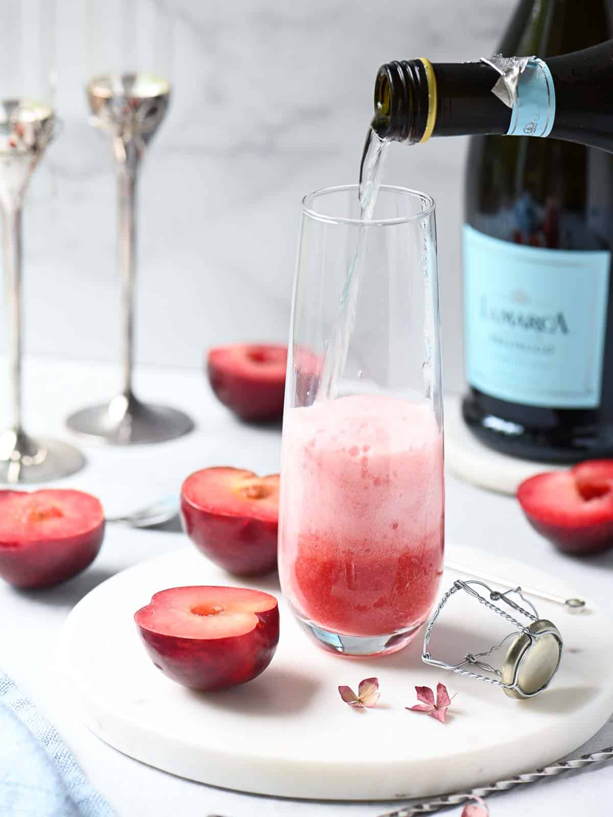 Prosecco being poured into a clear flute with fresh plums around it.