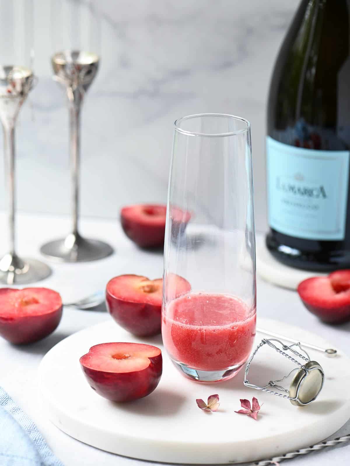 Plum puree in a clear flute with plums and a bottle of Prosecco in the background.