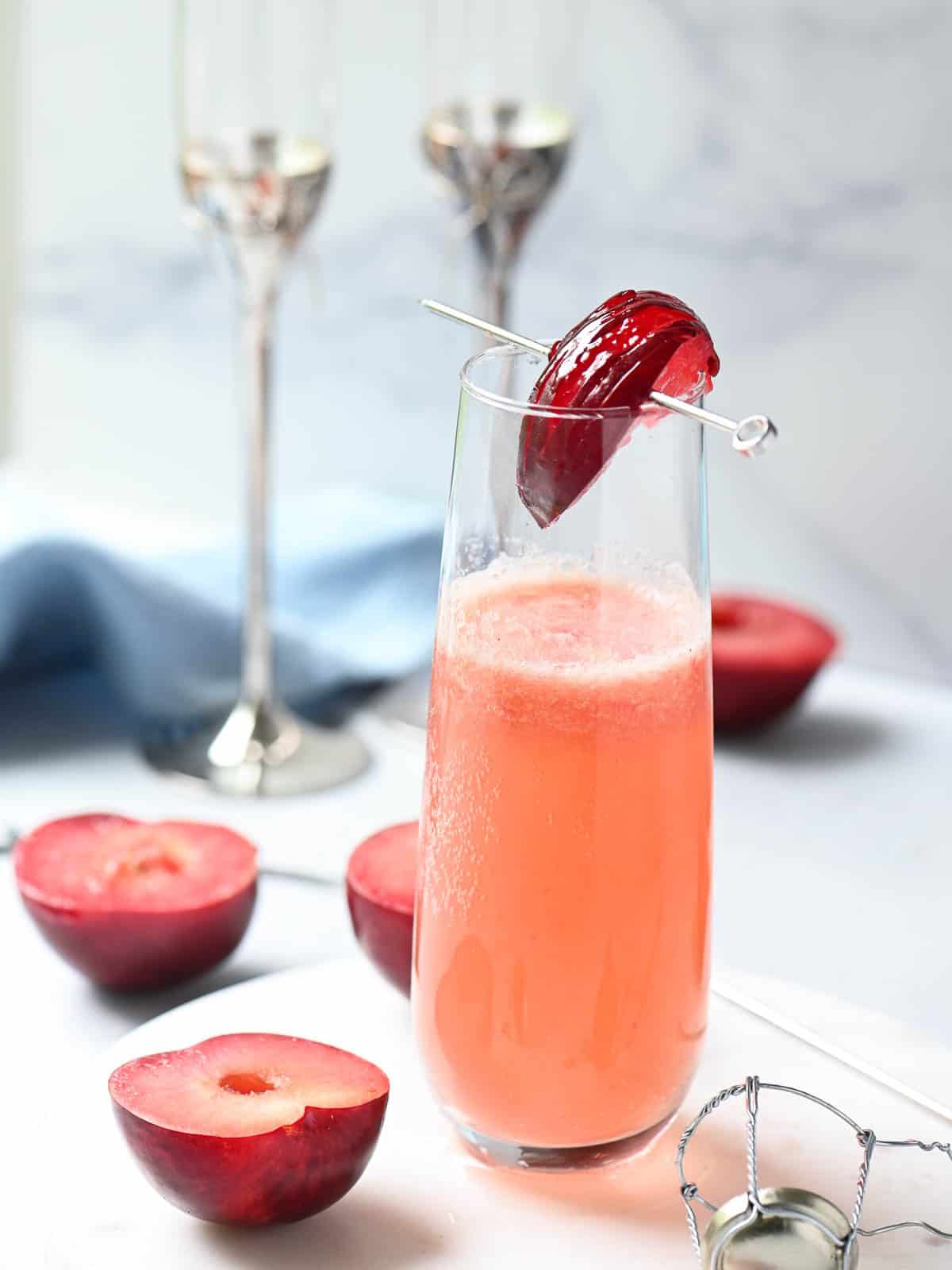 Plum bellini in a clear flute on a white surface with plums in the background.