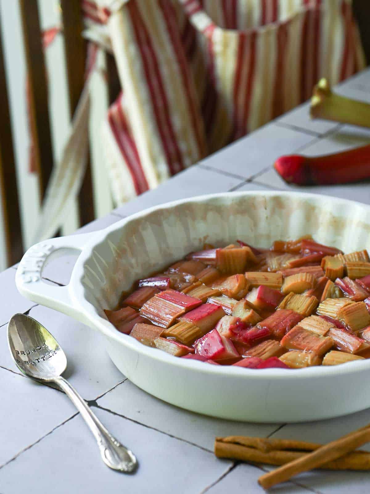 Roasted rhubarb in a white baking dish with a striped apron in the back.