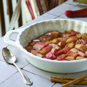 Roasted rhubarb in a white baking dish with a striped apron in the back.