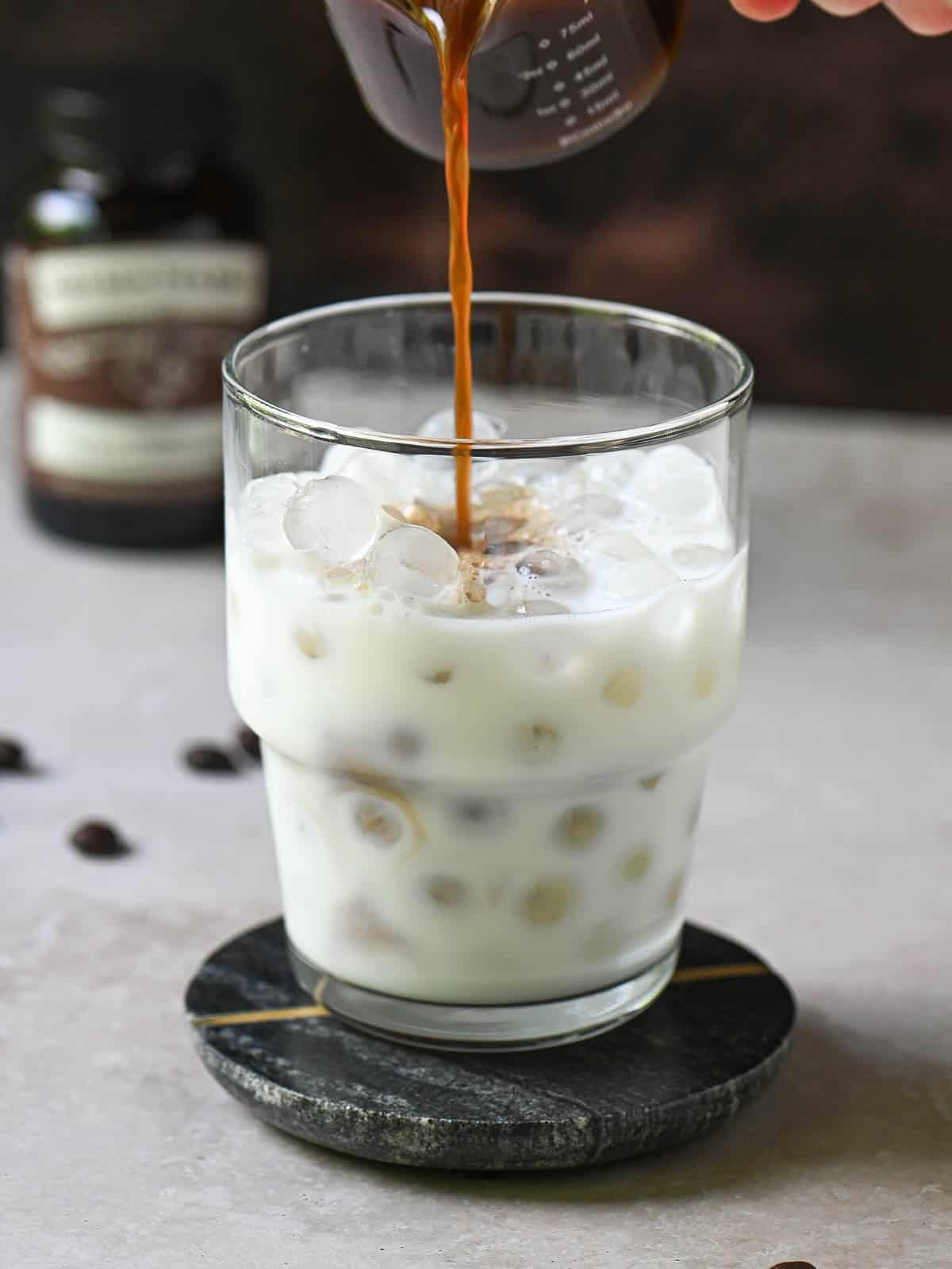 Coffee is being poured into a clear glass with milk and ice.
