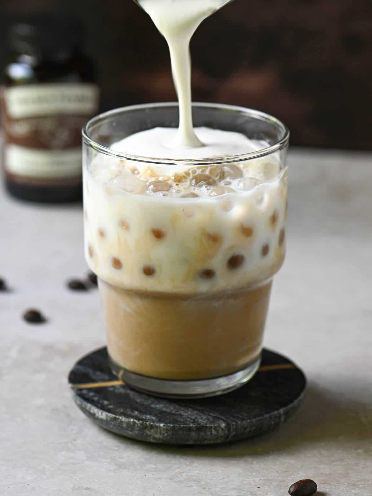 Cold foam is being poured into a clear glass with coffee and milk.